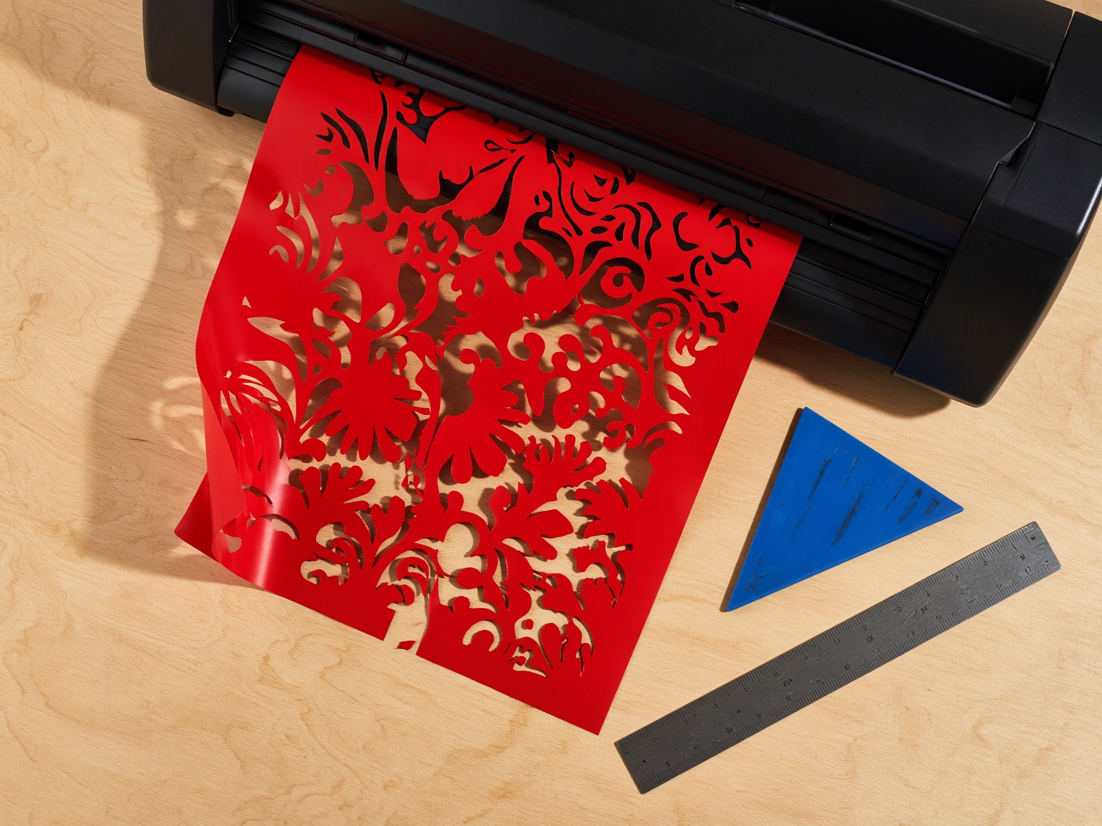 Red vinyl being contour-cut on a plotter beside a squeegee and steel ruler